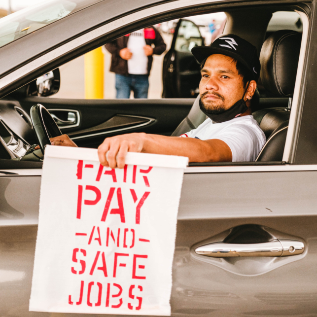 An app-based driver participates in an airport action. He holds a sign from his car that reads "Fair Pay and Safe Jobs."