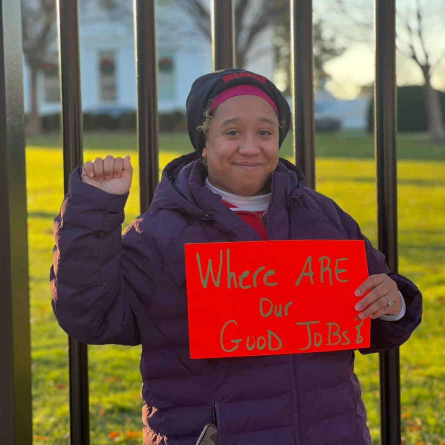 Black woman raising her fist and holding a sign that reads "Where are our good jobs?". She stands in a purple coat with a hoodie in front of the US Capitol gates.