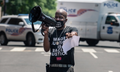 A Black man points directly at the photographer at an action. He is holding a bullhorn to the side and is wearing a mask that reads, "Fight, Don't Starve," and is wearing a shirt that reads "Wage Justice is Racial Justice."