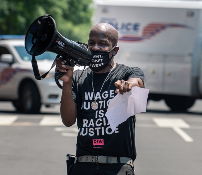 A Black man points directly at the photographer at an action. He is holding a bullhorn to the side and is wearing a mask that reads, 