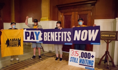 Four people from the Mon Valley Unemployed Committee stand outside the Governor's office. Three hold a sign that read, all together, "Pay Benefits Now." Another sign on the floor reads, "315,000 Still Waiting."