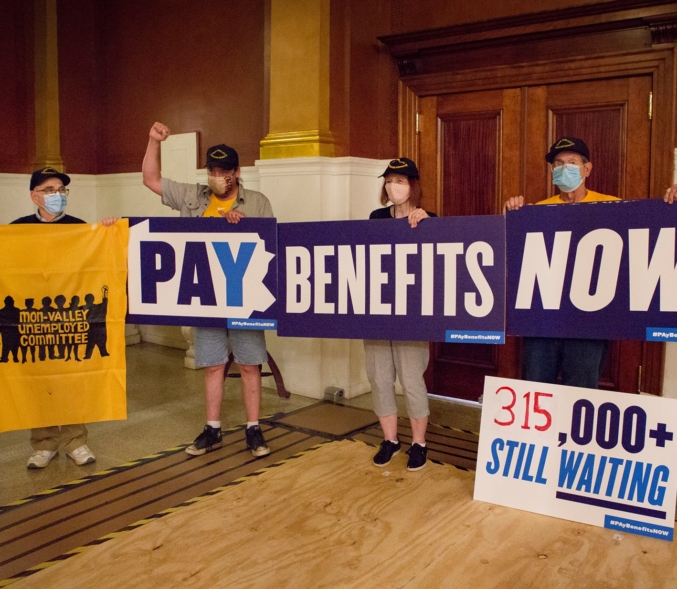 Four people from the Mon Valley Unemployed Committee stand outside the Governor's office. Three hold a sign that read, all together, "Pay Benefits Now." Another sign on the floor reads, "315,000 Still Waiting."