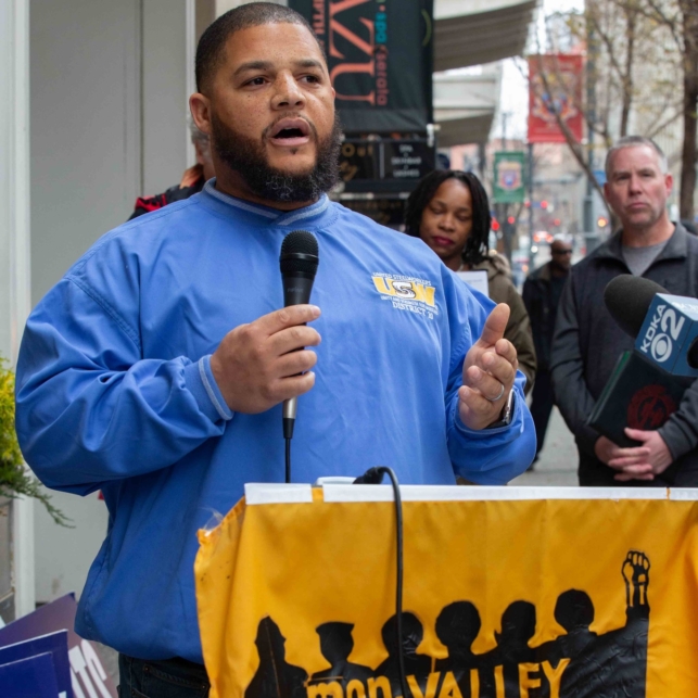 A Black man wearing a United Steelworkers shirt speaks into a microphone at an action with the Mon Valley Unemployed Committee.