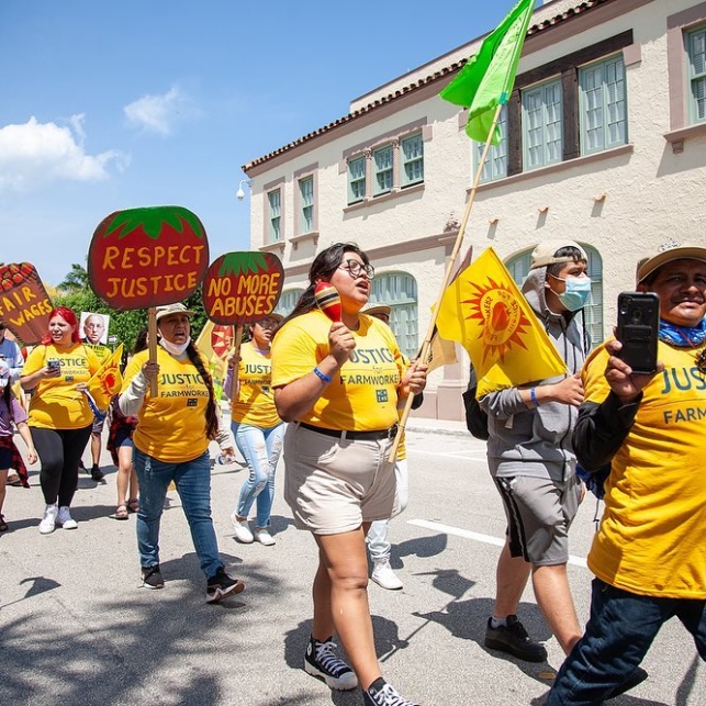 A group of workers pass by during an action. Their signs read, "Respect justice," "No more abuses," and "fair wages." They are wearing yellow "Justice for Farmworkers" shirts.