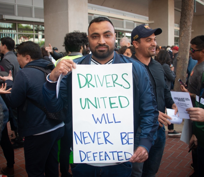 An app-based worker holds a sign that reads, "Drivers United Will Never Be Defeated."