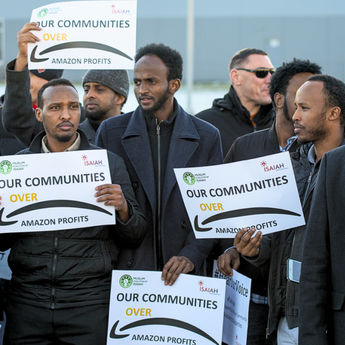 A group of men of color participate in an action. Their signs read, "Our Communities Over Amazon Profits."