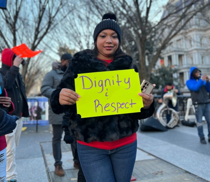 A Black woman holds a sign that reads "Dignity and Respect" at an action.