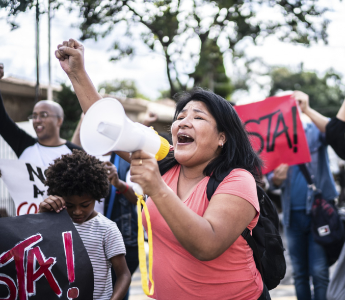 An AAPI woman speaking into a bullhorn leads a chant at an action.