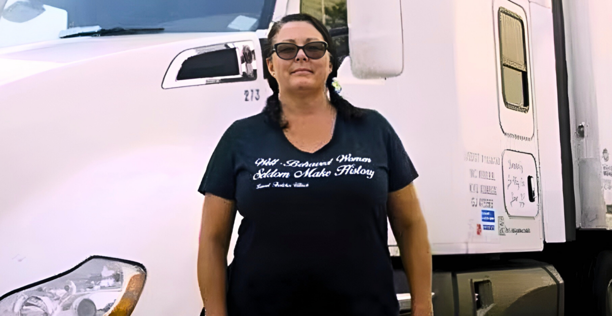 Desiree Wood, a truck driver, stands in front of her truck. She is wearing sunglasses and her shirt reads, "Well-Behaved Women Seldom Make History."