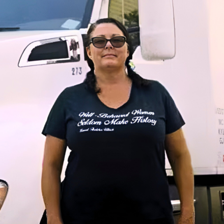 Desiree Wood, a truck driver, stands in front of her truck. She is wearing sunglasses and her shirt reads, "Well-Behaved Women Seldom Make History."