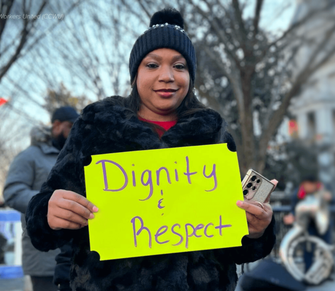 Black woman in black knit cap and coat standing outside in a protest holding a bright yellow handwritten sign that reads Dignity and Respect.