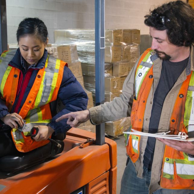 An industrial warehouse workplace safety topic. A safety supervisor or manager training a new employee on forklift safety. The trainer is holding a clipboard and explaining seat belt use to a younger female trainee.