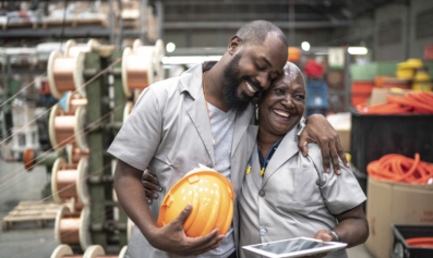 A Black construction worker holding a hat hugs his co-worker, a Black woman, who is holding a tablet computer.