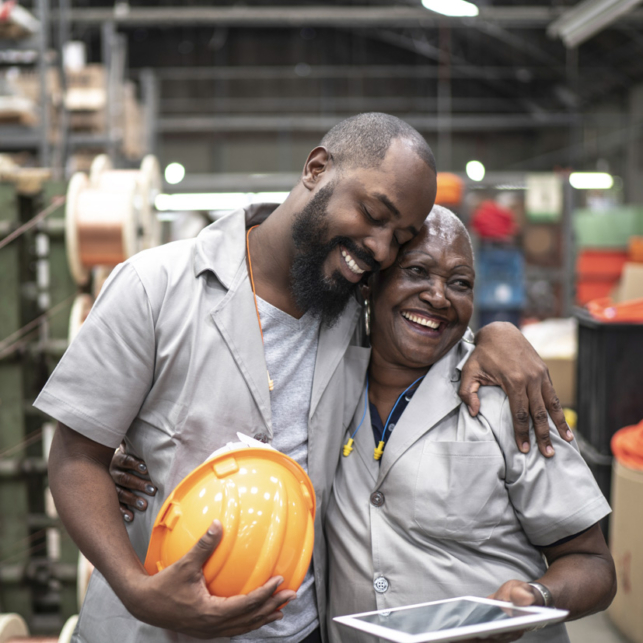 A Black construction worker holding a hat hugs his co-worker, a Black woman, who is holding a tablet computer.