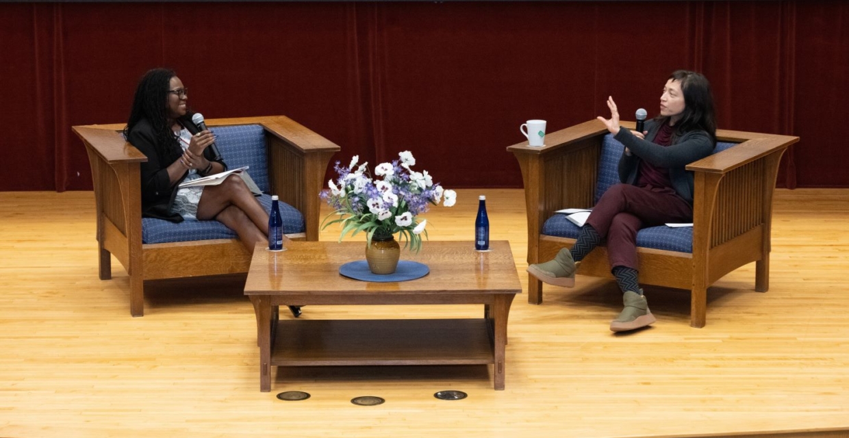 Two women seated on a stage in chairs hold microphones and talk to each other with a table between them. The table has a vase of purple and white flowers.