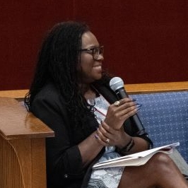 Two women seated on a stage in chairs hold microphones and talk to each other with a table between them. The table has a vase of purple and white flowers.