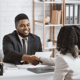 Image of a Black man smiling, shaking hands with a Black woman over a desk, with her facing toward him and away from the camera. They're in an office setting. They convey professionalism and camaraderie.