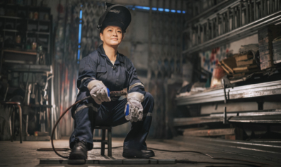An AAPI welder sits on a small stool in a warehouse. She has a calm, confident expression on her face.