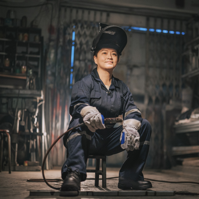 An AAPI welder sits on a small stool in a warehouse. She has a calm, confident expression on her face.