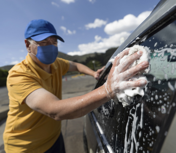 Adult Latin American man working at a car wash wearing a facemask while cleaning the windows of a car.