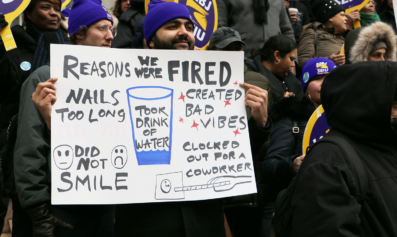 A worker at a City Hall action holds a sign that says "Reasons We Were Fired: Nails Too Long; Created Bad Vibes; Did Not Smile; Took Drink of Water; Clocked Out For A Coworker."