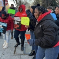 A group of 6 Black women in a circle holding picket signs and wearing coats.
