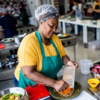 Middle-aged Black woman wearing a clear plastic safety cap, green apron and yellow short sleeved shirt shredding carrots in a kitchen.