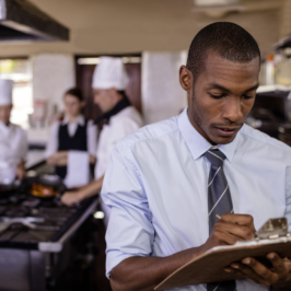 A young Black man writes on a clipboard with coworkers in a kitchen setting