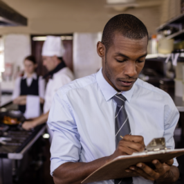 A young Black man writes on a clipboard with coworkers in a kitchen setting