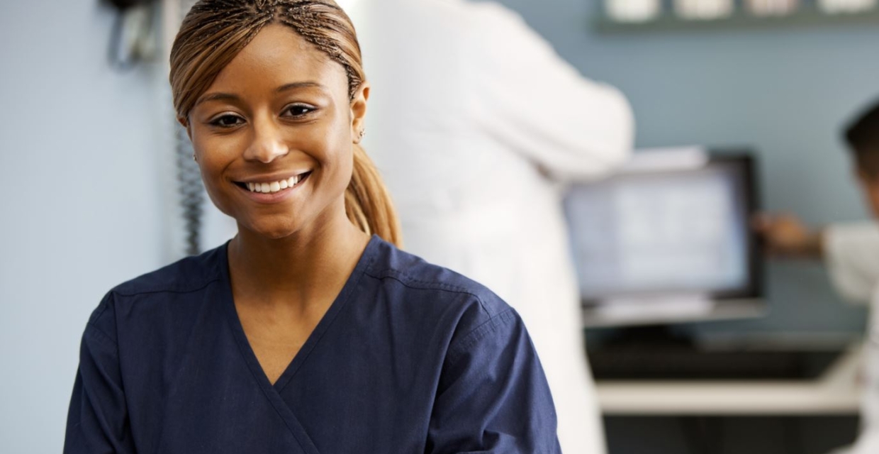 A young Black woman medical worker poses wearing navy blue scrubs with medical staff in the background
