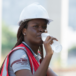 A Black woman worker wearing a white hardhat and red safety vest drinks a cold bottle of water outdoors