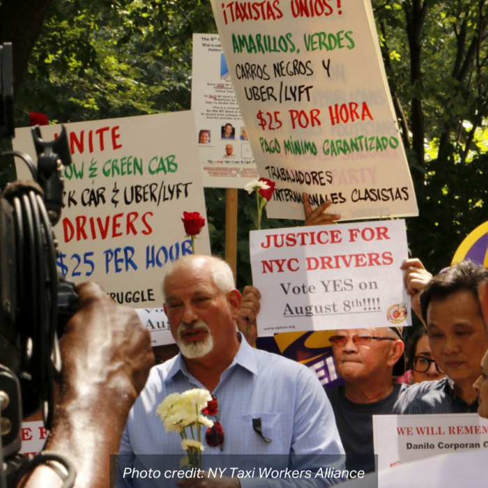A film crew records a Taxi driver holding white chrysanthemums in front of a rally of people holding signs that read: "Justice for NYC Drivers - Vote YES on August 8!", and "Taxistas Unios! - Amarillos, Verdes Carros Negros Y Uber/Lyft $25 Por Hora Pago Minimo Carantizado Trabajadores Internacionales Clasistas". photo credit: NY Taxi Workers Alliance