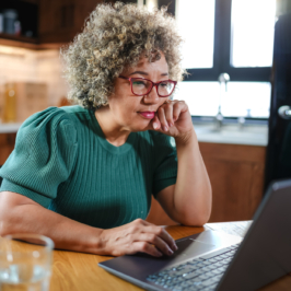 A concerned mature woman with curly gray hair reviews paperwork at her kitchen table. She holds a document while looking at it intently, surrounded by a laptop, calculator, and papers.