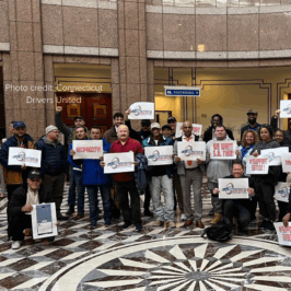 Advocates with Connecticut Drivers United stand in a municipal building carrying signs that read, "Transparency!" and "We Want S.B. 1180".