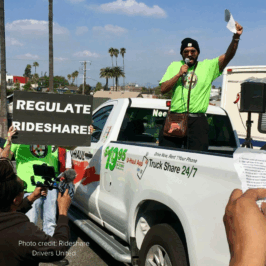 Advocates in bright green Rideshare Drivers United tshirts rally around a Uhaul pickup truck with a worker leader speaking into a microphone. A member carries a sign that reads, "Regulate Rideshare".