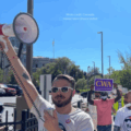 Image: Members of Colorado Independent Drivers United are participating in a protest. The person in the foreground on the left is holding a megaphone and wearing a white t-shirt with the logo “Colorado Independent Drivers” and “CWA” (Communications Workers of America). Several other protesters are visible behind him, some holding signs with messages such as “UBER + LYFT TAKE THE MAJORITY OF FARE.” The background shows trees, parked cars, streetlights, and buildings under a clear blue sky.