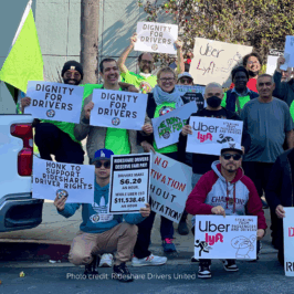 Image: Members of Rideshare Drivers United are gathered on a street corner near a white pickup truck, holding various protest signs advocating for better pay and rights, and in support of policies like the Empowering App-Based Workers Act. The signs include messages such as “Dignity for Drivers,” “Fair Pay Now,” “No Deactivation Without Representation,” and critiques of Uber and Lyft’s pay practices. One sign highlights that drivers make $6.20 an hour while the Uber CEO makes $11,538.46 an hour. Another sign opposes Proposition 22 with a red circle and slash over “Prop 22.” The setting is outdoors on a sunny day.