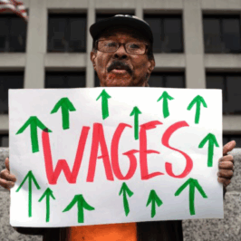 A Black man wearing a navy baseball cap and glasses in front of a municipal building holds a poster that reads, "WAGES" with upward green arrows. Raise the MInimum Wage, Living Wage, Raises, Increase minimum wage, Fair Wage movement,
