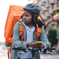 A Black woman worker at an app-based delivery job sits on a bike while carrying a large orange pack and holding a mobile phone.