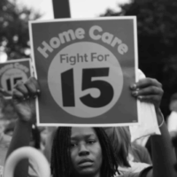A black and white image of an African American woman amid a crowd carries a sign reading "Home Care Fight for 15".