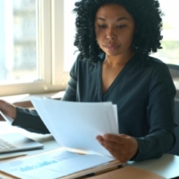 A Black woman in a black blouse looks over paperwork at her desk in an office.