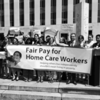 A photo of a diverse group of people standing on steps outside a large building, holding signs and a large banner advocating for fair pay for home care workers, which would include minimum wage and overtime protections. The prominent banner reads, “Fair Pay for Home Care Workers - Helping others live independently shouldn’t mean living in poverty.” Many individuals hold smaller signs with similar messages supporting home care workers and family caregivers. The group appears united in a peaceful protest or rally to raise awareness about the need for better wages and support for home care workers.