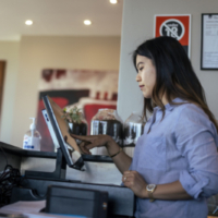 A tipped-worker stands behind the bar of a restaurant and uses a touch screen computer to take an order.