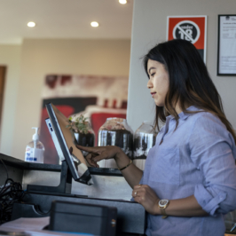 A tipped-worker stands behind the bar of a restaurant and uses a touch screen computer to take an order.