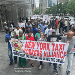 Group of app-based drivers and supporters march in Manhattan holding a banner reading “NEW YORK TAXI WORKERS ALLIANCE” and placards demanding respect, fair pay, and protections for drivers, which would include just cause / deactivation protections. Skyscrapers and city streets frame the organized rally.