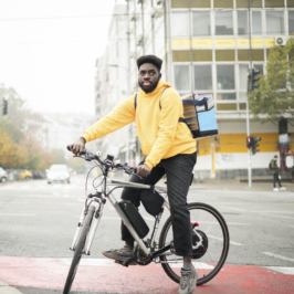 A young Black courier carries a delivery box on his back while riding a bike in the city.