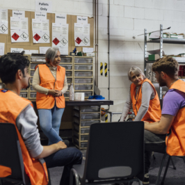 A multiracial group of five workers wearing orange safety vests have a discussion seated in a circle in a warehouse breakroom.