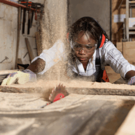 A young Black woman wears hearing protection and gloves as she works a table saw in a manufacturing job.