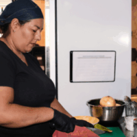 A middle-aged Latina woman chops jalapenos in a restaurant kitchen. She wears a denim bonnet, black t-shirt, and black latex gloves.