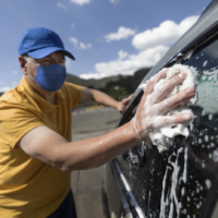 Adult Latin American man working at a car wash wearing a facemask while cleaning the windows of a car.